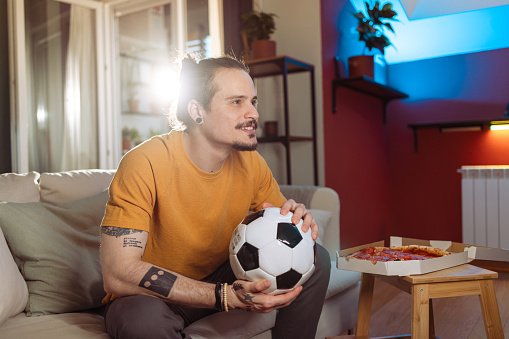 Photo of a young Latin man sitting on the couch in the living room and holding a sports ball while watching a football game. A Latin American man is wishing for a goal.