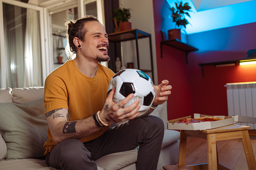 Photo of a cheerful Latin American man sitting on the couch in the living room and cheering. He is celebrating a goal that his favourite club achieved.