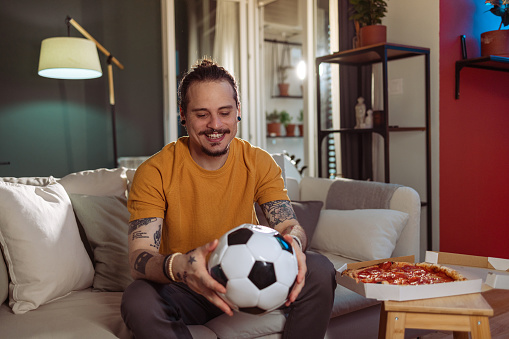 Latin-American man is ready to watch a soccer game and to support his favourite team. He is holding a ball and ordered a delicious take-out pizza.
