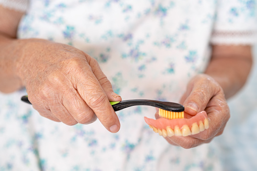 Asian elderly woman patient use toothbrush to clean partial denture of replacement teeth.