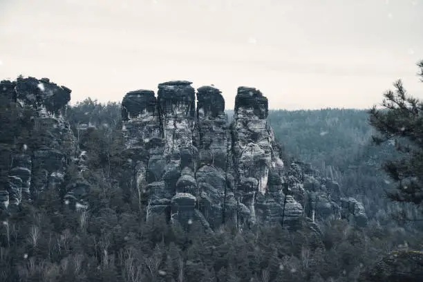 Rugged rocks at Basteibridge during snowfall. Wide view over trees and mountains. National park in Germany