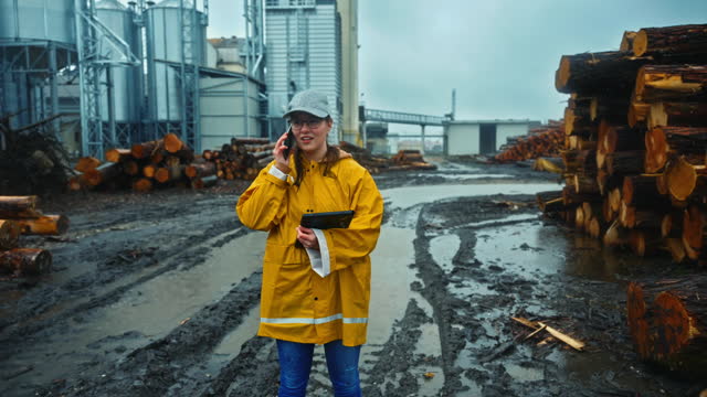SLO MO Young Female Worker in Raincoat and Cap Talking on Smartphone at Lumberyard During Rainy Season