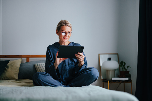 Close up shot of a beautiful smiling senior woman sitting on the bed in her pajamas. She is wearing glasses, holding and using digital tablet. She is looking down and smiling.