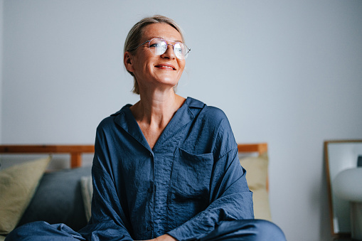 Close up shot of a beautiful older woman wearing glasses and sitting on the bed in her pajamas enjoying the morning at home. She is smiling and looking away.