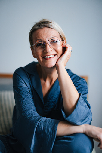 Close up shot of a beautiful older woman wearing glasses and sitting on the bed in her pajamas enjoying the morning at home. She is smiling and looking at the camera.