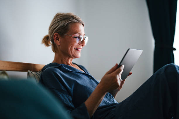 Portrait of a Senior Woman Relaxing in Her Bed and Using Digital Tablet Close up shot of an older woman sitting and relaxing in her pajamas in the bed at home. It is the morning and she is using a digital tablet to go on the internet, or have a video call, or watch something. She is smiling while looking down. old woman smiling tablet stock pictures, royalty-free photos & images