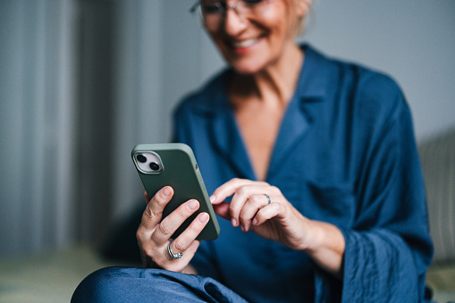Close up of an unrecognizable senior woman sitting in her pajamas and holding mobile phone. She is using it for texting or reading a message.