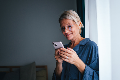 Close up shot of a senior woman wearing glasses and holding mobile phone. She is smiling while looking down and texting in the morning at home.
