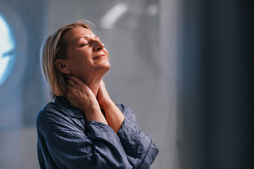 Close up shot of a beautiful older woman standing in her pajamas in the bathroom at home. She is either massaging her neck or applying face cream on her neck and face. She has her eyes closed.