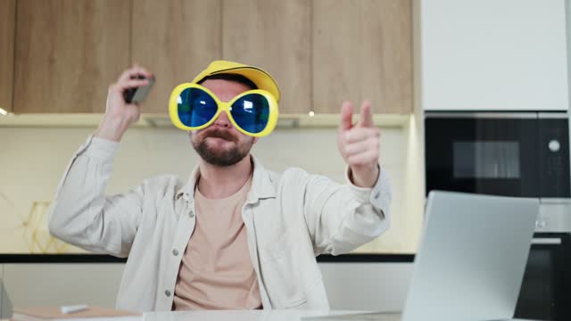 Caucasian man in funny eyewear and yellow headdress holding smartphone and celebrating victory in apartment. Excited gadget user waving hands with clenched fists and pointing fingers at camera.