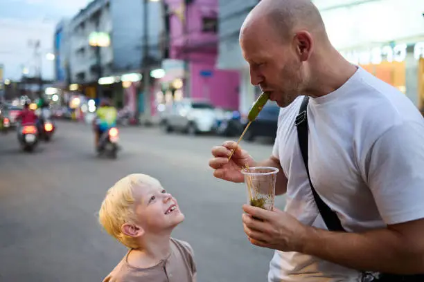 A father tastes food with his 6 year old son at a night market in Thailand during their vacation in Asia