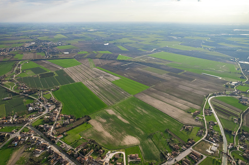 The aerial view shows a vast expanse of green field in the Pianura Padana region. The field is dotted with patches of various shades of green, indicating different crops or grass types. The field extends to the horizon, with no visible boundaries or obstacles.