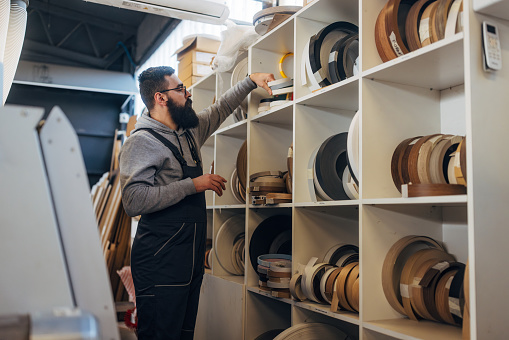 A bearded craftsman in a uniform is standing at a shelf with edging tapes and choosing which he needs when making furniture