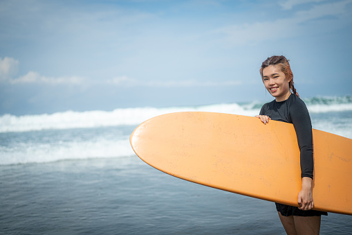 Radiant Asian surfer stands confidently by the ocean, joyfully embracing her new hobby. With a captivating smile and chic black swimsuit, she embodies the essence of wellness travel.