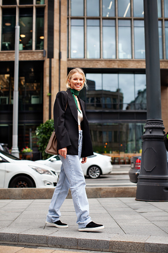Beautiful young stylish woman walking down city street against backdrop of building, wearing fashionable clothes jeans, black blazer, handbag and sneakers. Happy Fashion model.