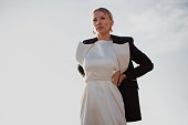 Woman Fashion model strikes a pose on sky background, adorned in luxurious attire long white dress and a black blazer, showcasing a stylish ensemble