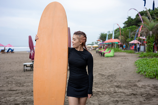 Radiant Asian surfer stands confidently by the ocean, joyfully embracing her new hobby. With a captivating smile and chic black swimsuit, she embodies the essence of wellness travel.