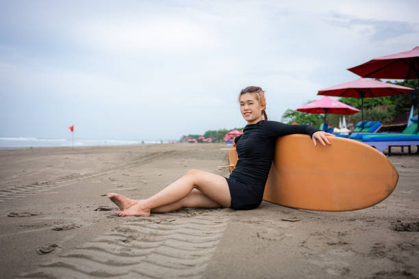 A beautiful young Asian woman relaxing on the beach with her surfboard. Looking at camera