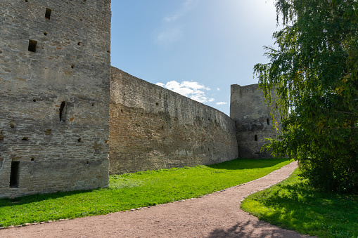 Walls of an old Russian fortress made of local stone with traces of time and destruction, watchtowers with embrasures, powerful defensive structures, fortification architecture, history and culture, city landmark, green trees and lawns, autumn view.