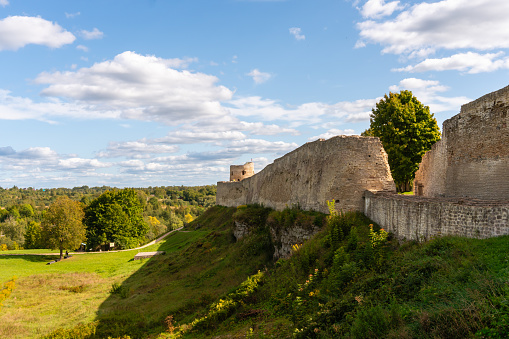 The walls of an old Russian fortress made of local stone with traces of time and destruction, watchtowers with embrasures, powerful defensive structures, fortification architecture, an earthen rampart and the surrounding area, history and culture, a city landmark, green trees and lawns, an autumn view.