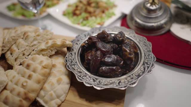 Vintage Islamic style metal bowl filled with dates, accompanied by flatbread, water, chicken salad, and other food items, arranged on a table for Muslims to break their fast during Ramadan