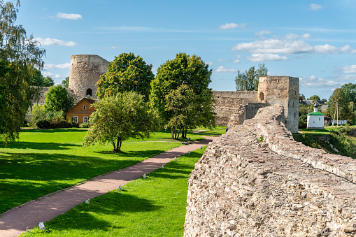Walls of an old Russian fortress made of local stone with traces of time and destruction, watchtowers with embrasures, powerful defensive structures, fortification architecture, wooden house, history and culture, city landmark, green trees and lawns, autumn view.