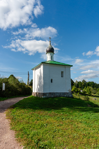 A small simple chapel in the old Russian style with a dome and a cross near the walls of an old fortress, thick walls and embrasures, an Orthodox church, green lawns and trees, sky and clouds, history and culture, travel and tourism, autumn.