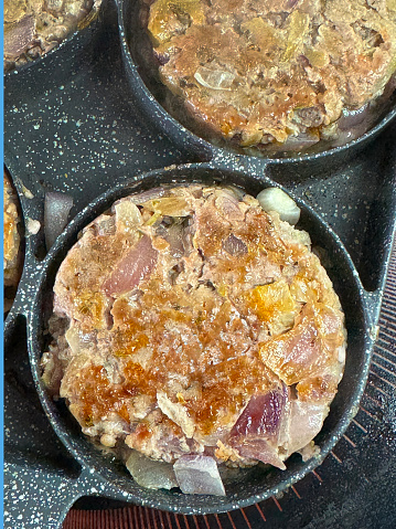 Stock photo showing close-up view of four fried sausage meat and red onion burger patties that are being cooked in a stainless steel, non-stick frying pan on a ceramic hob. The pan has rings to keep the contents separate and stop them from merging.