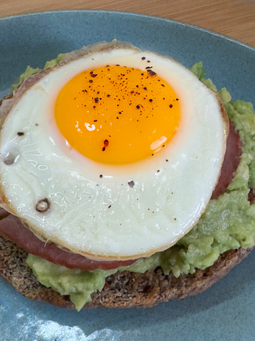 Stock photo showing close-up, elevated view of a green-blue plate containing a wholemeal bagel topped with mashed avocado, ham and a sunny-side-up fried egg, in a restaurant setting.