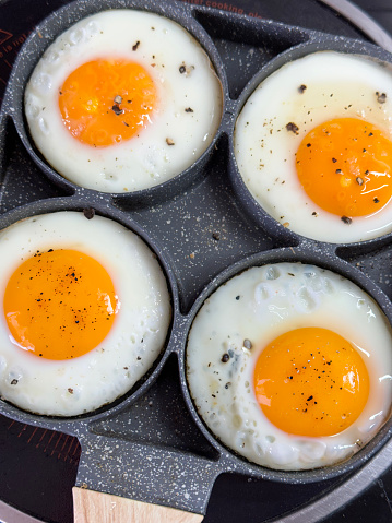Stock photo showing close-up view of four sunny-side-up, fried eggs that are being cooked in a stainless steel, non-stick frying pan on a ceramic hob. The pan has rings to crack eggs into in order to keep them separate and stop the egg whites from merging.