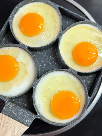 Stock photo showing close-up view of four fried eggs that are being cooked in a stainless steel, non-stick frying pan on a ceramic hob. The pan has rings to crack eggs into in order to keep them separate and stop the egg whites from merging.