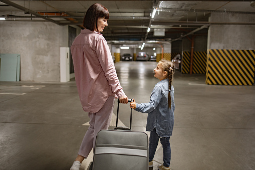 Attractive lady with her child and travel suitcase, searches for their car before starting trip. Dressed in informal attire, Caucasian mom and small daughter walking through underground parking lot.