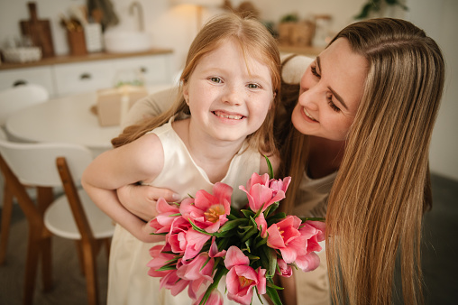 Mom and kid hugging, smiling. Happy family holiday. Cute little child girl greeting mother in kitchen. Beautiful daughter gives mother big bouquet of tulip flowers and makes surprise for Mother's Day.