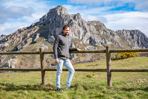 A middle aged man leaning on a fence in a meadow with the mountains at his back in a sunny day