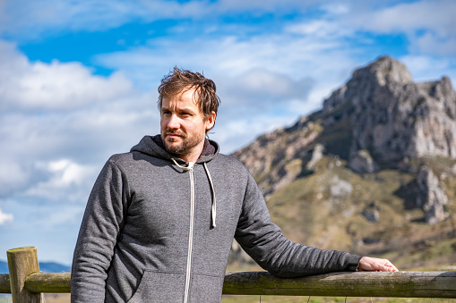 A portrait of a middle aged man leaning on a fence in a meadow with the mountains at his back in a sunny day