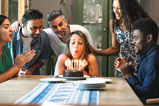 young woman blowing out candles on birthday cake surrounded by friends