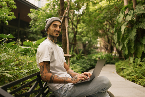 Happy hipster man using computer while relaxing on a bench in nature and looking at camera.