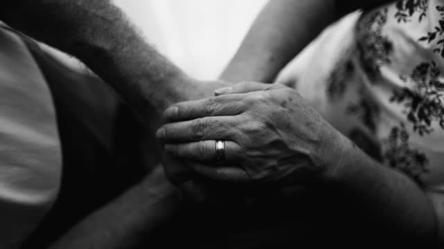 Elderly couple's hands in caring gesture of support and help during challenging times, seen from above of wrinkled hand on top of each other in black and white