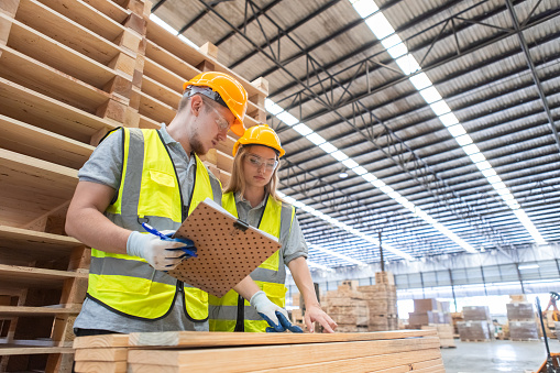 Team engineer carpenter wearing safety uniform and hard hat working holding clipboard checking quality of wooden products at workshop manufacturing. man and woman worker wood warehouse industry.