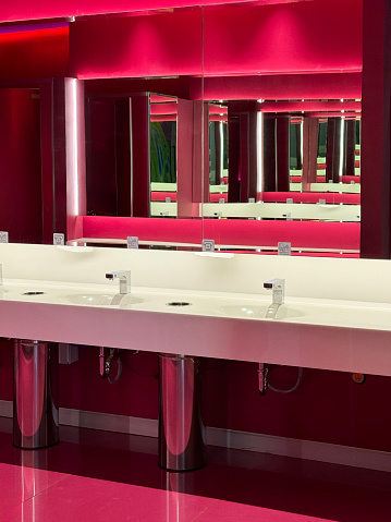 Stock photo showing red public toilet washroom with row of white, bathroom counter inset sinks with infrared basin mixer taps in front of large wall mounted mirror that is reflecting a row of red, toilet cubicle doors.
