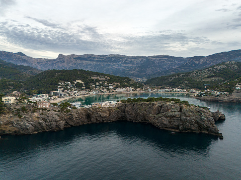 The aerial view at dusk of Port de Soller, located in Mallorca, Spain, captures the serene beauty of the harbor as the sun sets over the sea. The image showcases the charming town of Port de Soller, with boats dotting the harbor and lighthouses standing on the right and left.