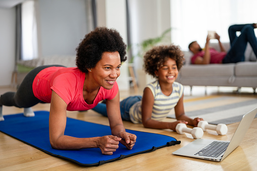 Happy young african american woman with child watching online video sports training and stretching on fitness mat. Concept of family, healthcare, sports and yoga at home