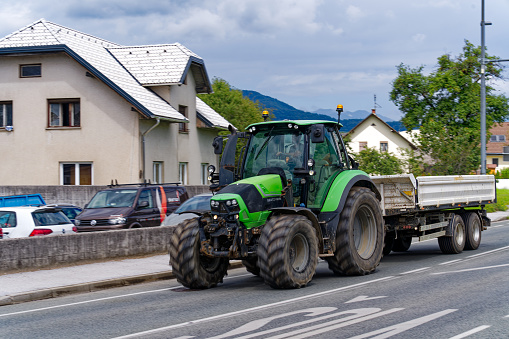 Green tractor with trailer passing on rural road at Slovenian village of Zabnica on a cloudy summer day. Photo taken August 10th, 2023, Žabnica, Slovenia.