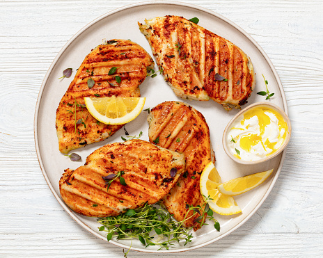 boneless skinless juicy grilled chicken cutlets with yogurt sauce and lemon slices on plate on white wooden table, horizontal view from above, flat lay, close-up