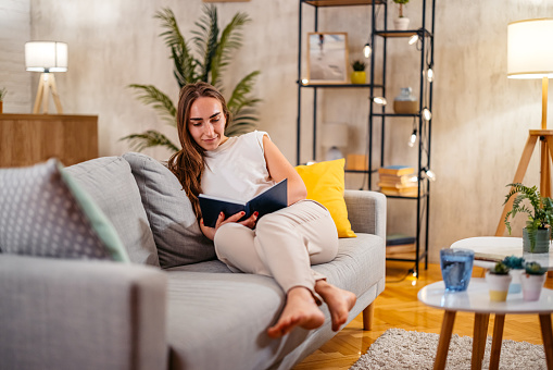 Beautiful young woman sitting on the sofa at home and reading a book.