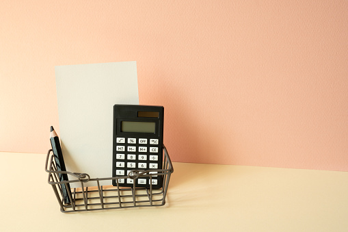Consumer shopping basket with calculator, pencil, memo pad on ivory and pink background. Economic Concept