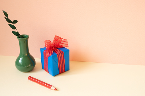 Blue gift box, vase of eucalyptus leaf, red colored pencil on ivory table. pink wall background