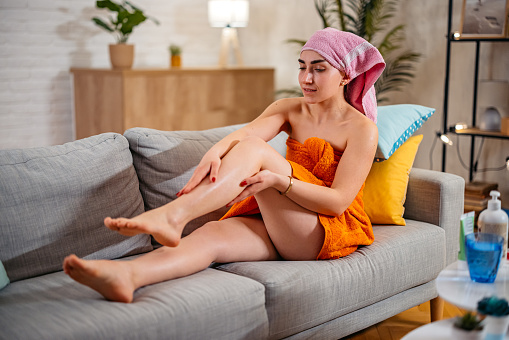 Beautiful young woman in a towel applying moisturizer on her legs while sitting on the sofa in the living room.