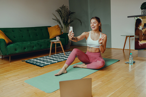 A woman is sitting comfortably on a plantbased yoga mat in her living room, taking a selfie