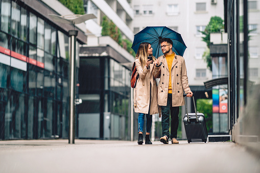 a beautiful young couple of tourists, walking in the rain with a suitcase and a large umbrella